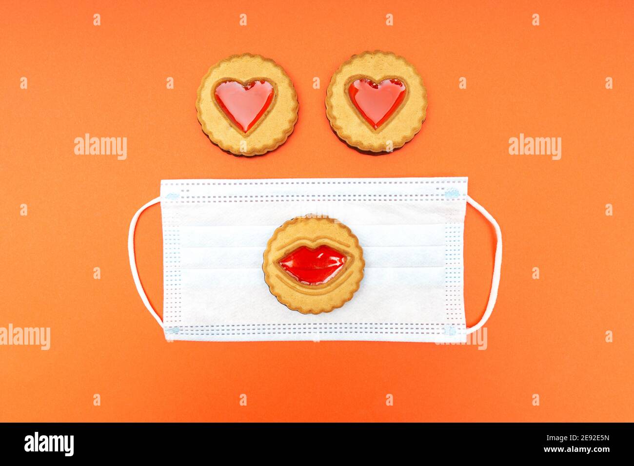 Lustige Figur aus einer Einweg-Gesichtsmaske und Kekse mit Lippen und Herzen auf einem orangen Hintergrund. Valentinstag Feier während der Quarantäne Stockfoto