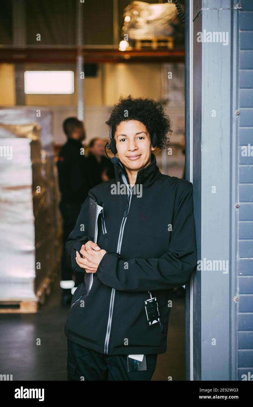Selbstbewusste manuelle Mitarbeiterin, die an der Wand im Logistiklager gelehnt ist Stockfoto