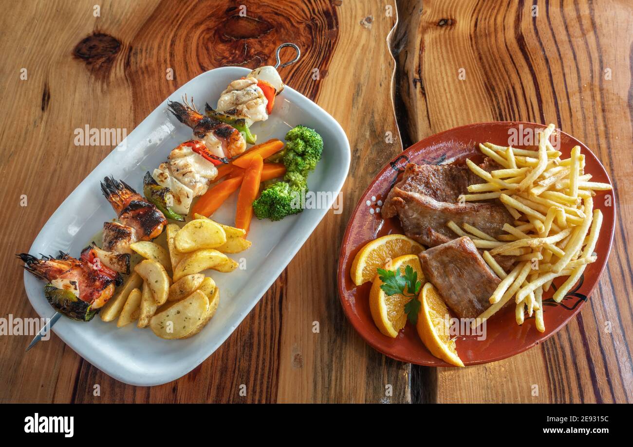 Gegrillter Fisch-Spieß mit Pommes und Gemüse auf der linken Seite. Teller Steak mit Pommes frites, garniert mit Orange auf der rechten Seite. Stockfoto