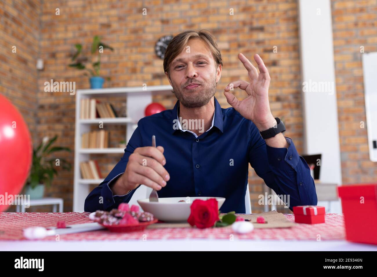 Lächelnder kaukasischer Mann, der einen Videoanruf beim Essen am Tisch macht Eine „OK“-Geste Stockfoto
