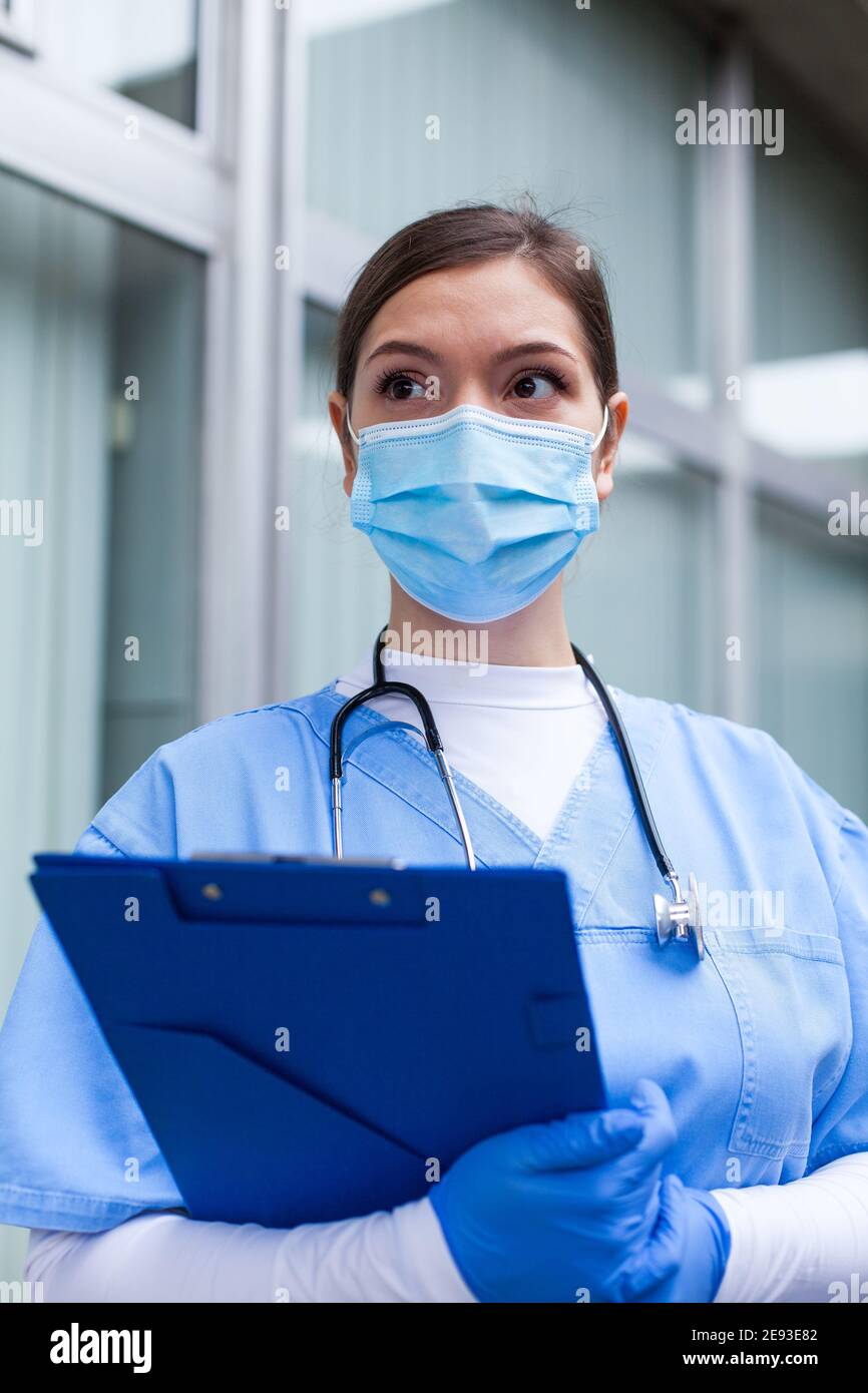 Junge weibliche kaukasische UK NHS GP Arzt suchen in der Ferne Mit Angst, Sorge, Angst in den Augen, trägt blaue ICU einheitliche Gesichtsmaske, Hoffnung und Vertrauen in overc Stockfoto