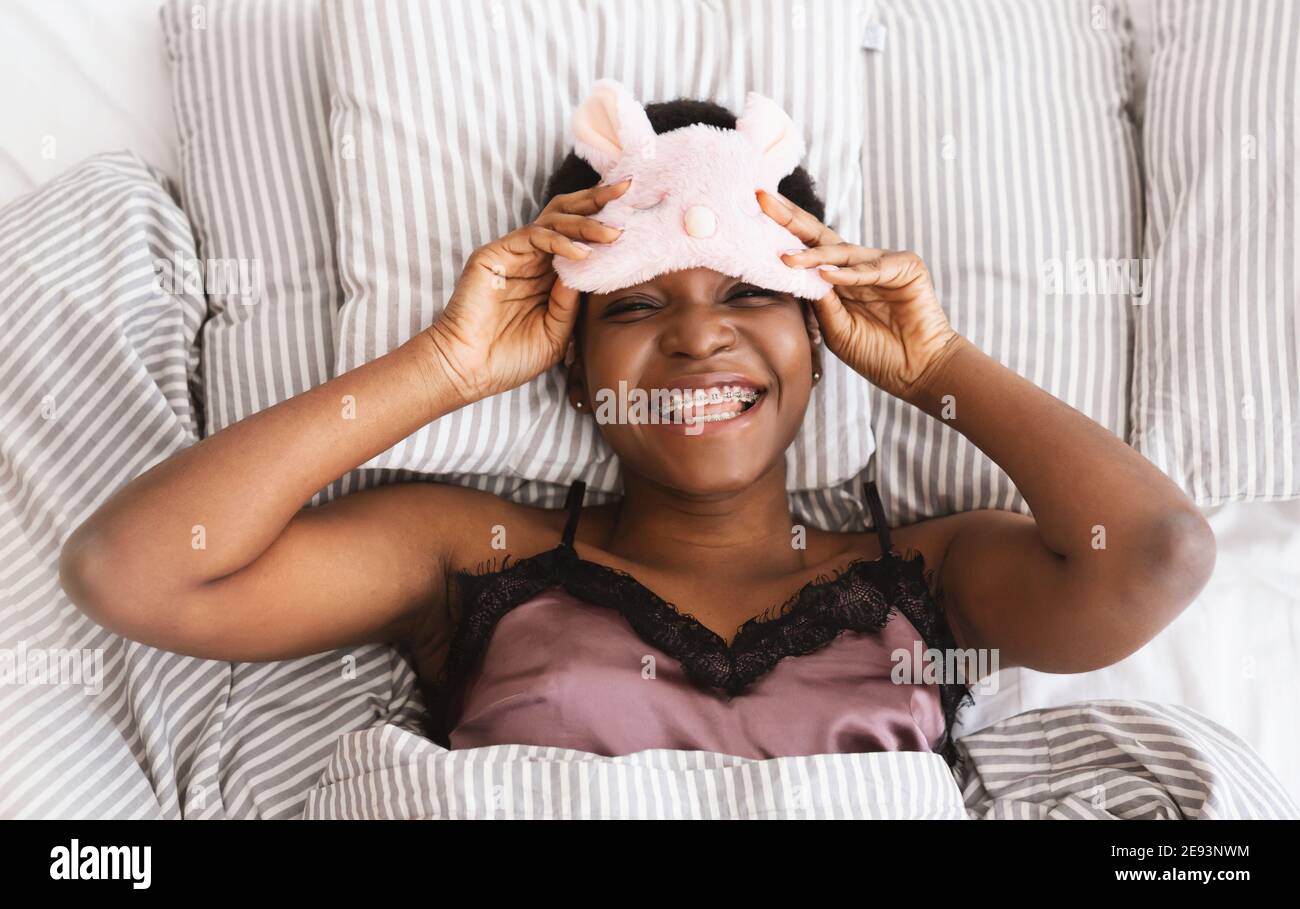 Faul fröhlich Millennial afroamerikanische Frau zieht aus Schlafmaske und schaut in die Kamera, liegt auf dem Bett Stockfoto