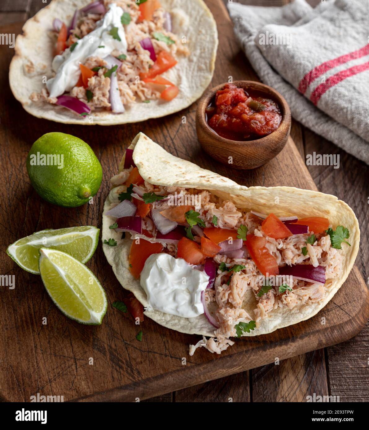 Taco mit gezogenem Huhn, Tomate, Zwiebel und saurer Sahne mit Limettenkeilen und Salsa an der Seite auf einem Holzbrett Stockfoto