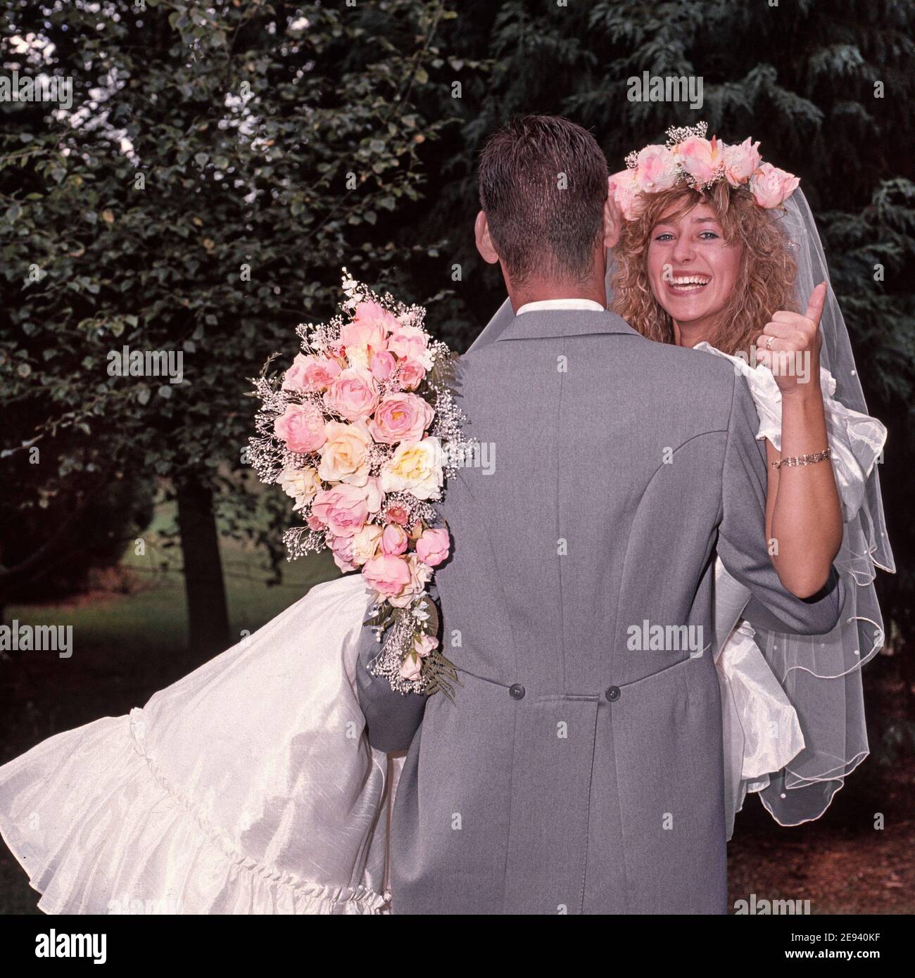 Hochzeit im Jahr 1989 von jungen Modell freigelassene Paar posiert im Freien Für Fotografien in 80s Mode Braut im Hochzeitskleid mit Blumenstrauß gibt Daumen nach oben getragen vom Bräutigam Rückansicht Grauer Morgenanzug und das Tragen einer lächelnden glücklichen Braut historischen 1980s Archiv Essex England Großbritannien Stockfoto
