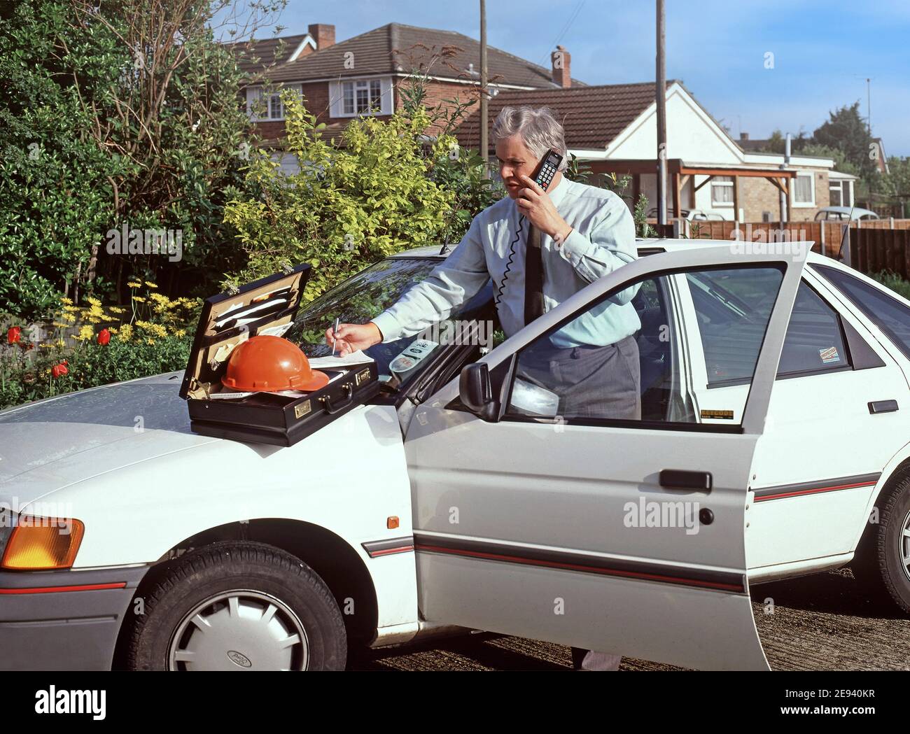 Bild eines Geschäftsmannes, eines Bauleiters, der an einem frühen, kabelgebundenen Mobiltelefon von der offenen Tür eines Ford Escort Firmenwagens spricht, historisches Archiv der 1990er Jahre Essex England Großbritannien Stockfoto