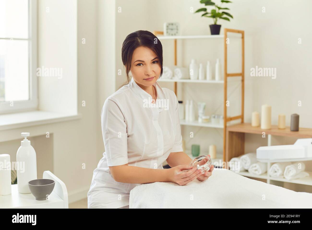 Lächelnde Frau Dermatologe sitzt, hält feuchtigkeitsspendende Maske im Glas und Blick auf die Kamera Stockfoto