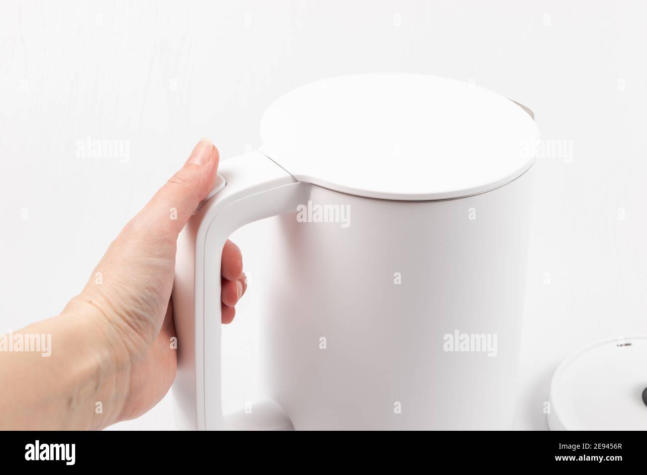 Eine weibliche Hand hält einen weißen Wasserkocher an der Griff auf einem weißen Holztisch Hintergrund Stockfoto