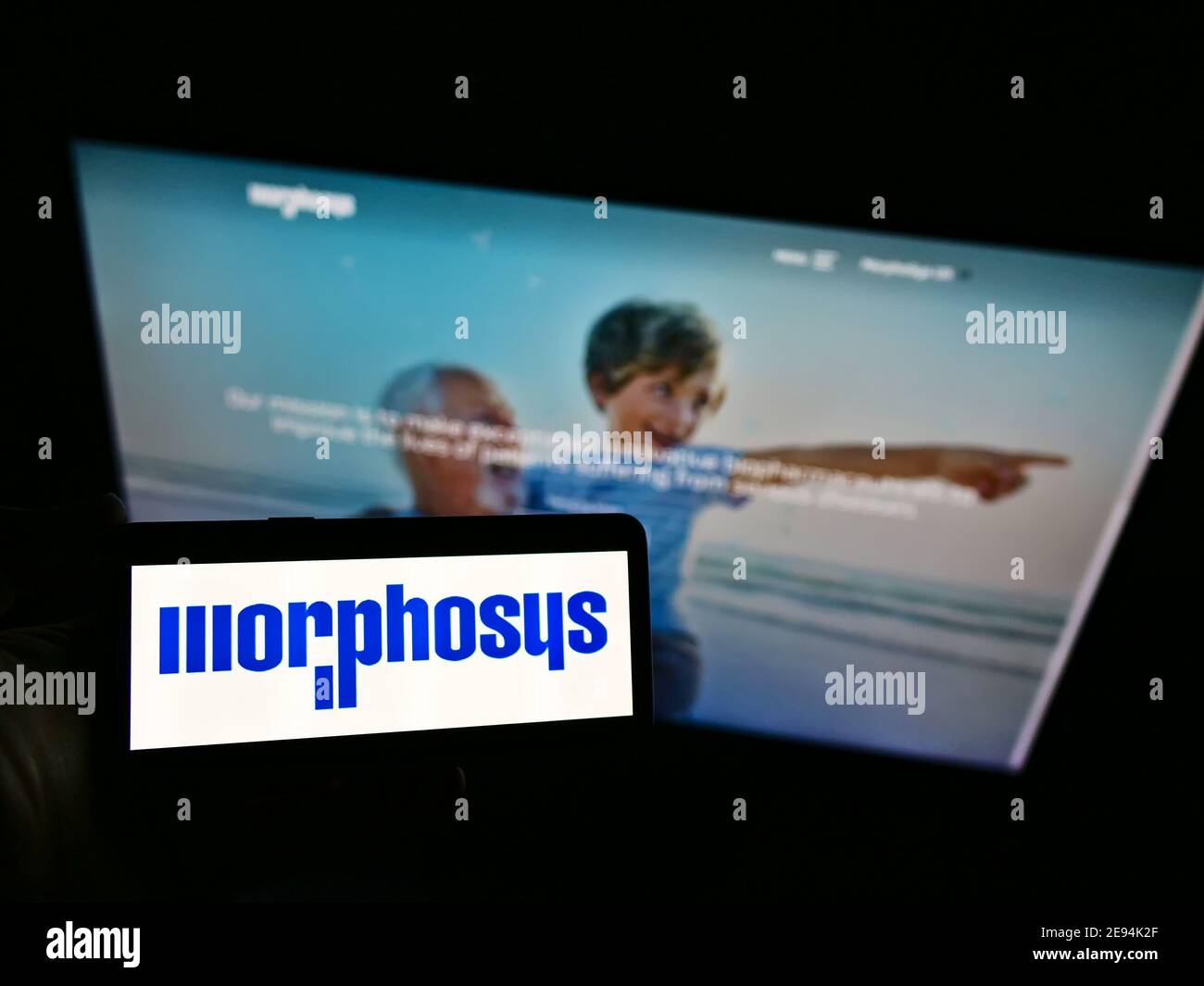 Hochwinkelansicht der Person, die Mobiltelefon mit Logo des deutschen Biotechnologie-Unternehmens MorphoSys AG hält, auf dem Display mit Website. Fokus auf Telefonbildschirm. Stockfoto