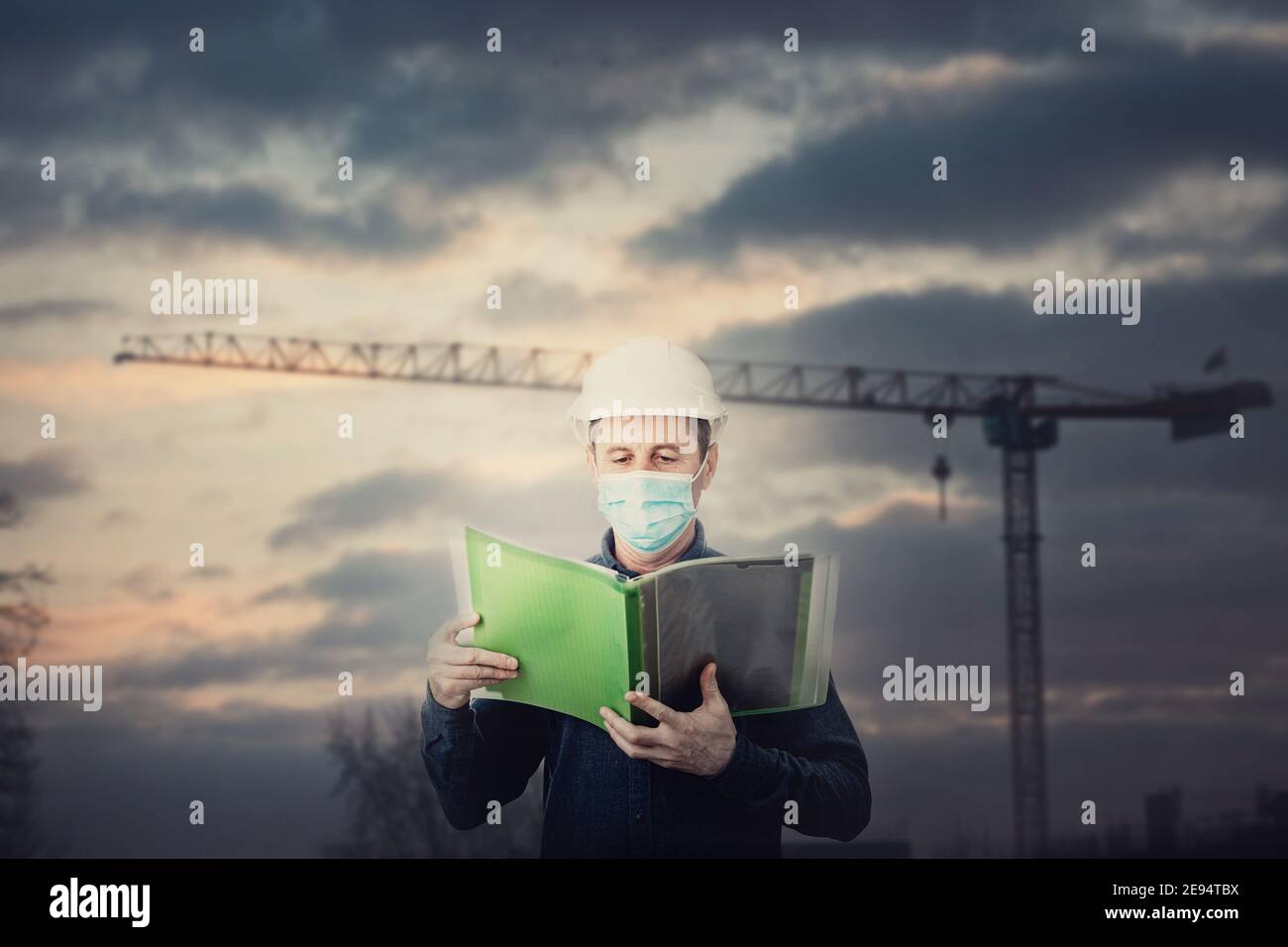 Auftragnehmer oder Architekt mittleren Alters, der eine Dokumentenmappe hält, Schutzhelm und Gesichtsmaske als Covid-19-Prävention trägt. Ingenieur mit Hardhat o Stockfoto