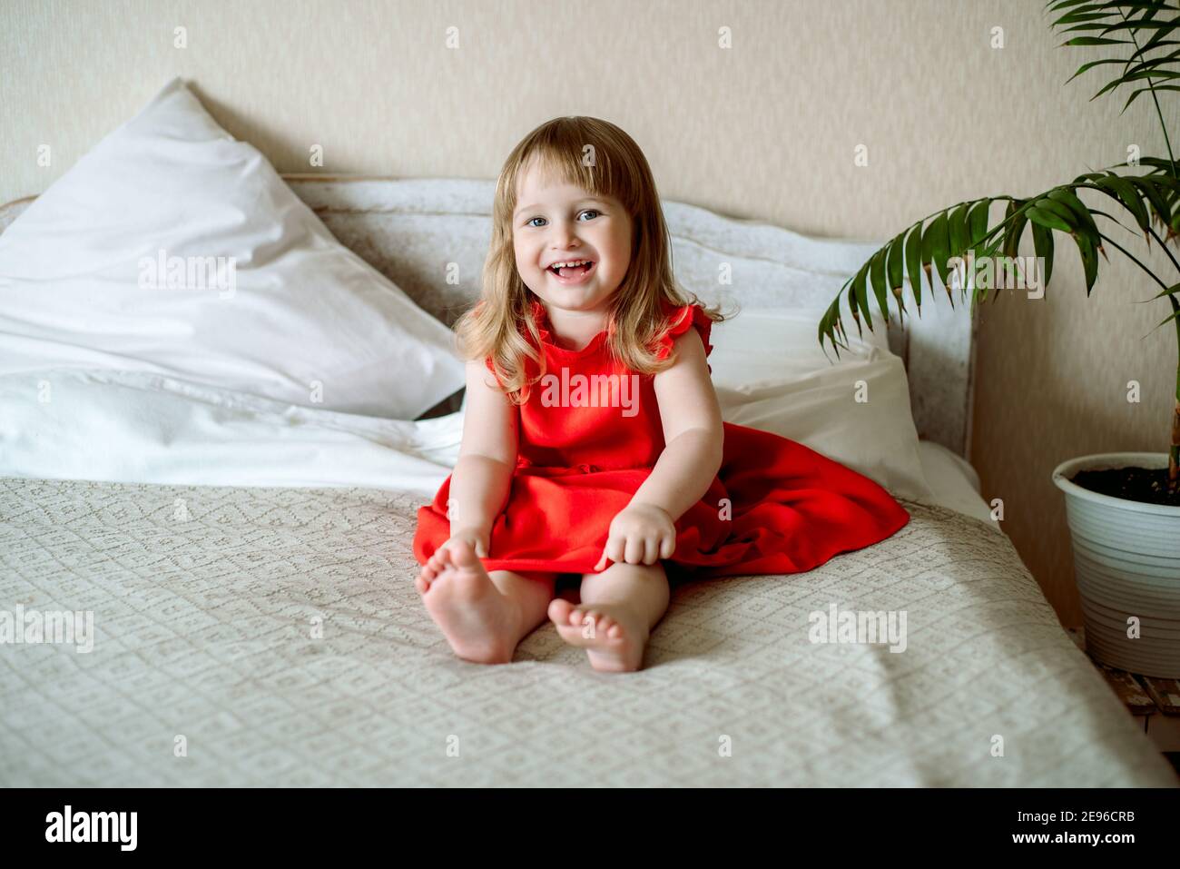 Niedliches rothaariges blauäugiges emotionales Mädchen lacht. Liegt auf dem Bett im Schlafzimmer, das Bett ist ein weißes Bett, ein rotes Kleid für ein Kind. Kissen und Stockfoto