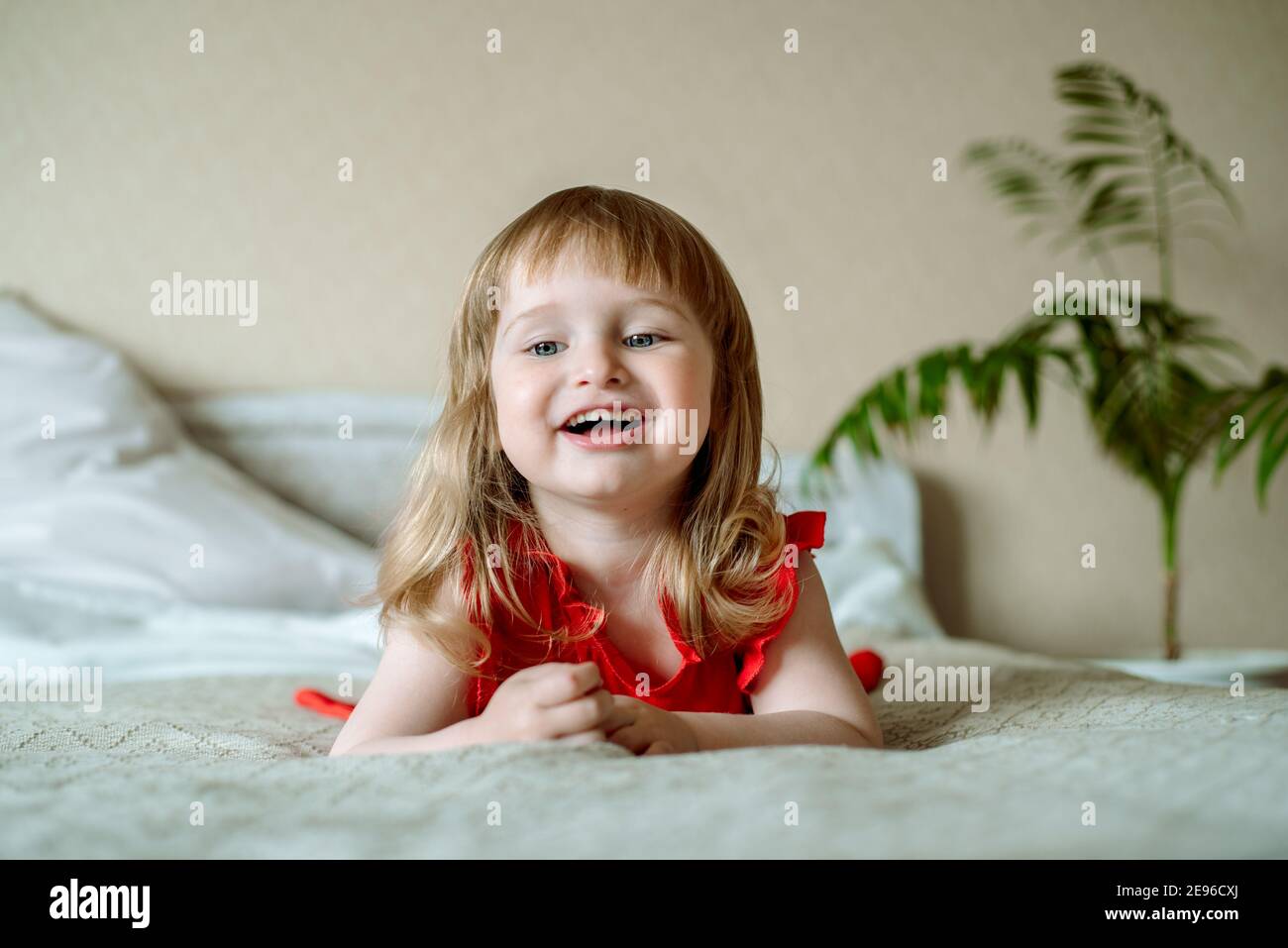 Niedliches rothaariges blauäugiges emotionales Mädchen lacht. Liegt auf dem Bett im Schlafzimmer, das Bett ist ein weißes Bett, ein rotes Kleid für ein Kind. Kissen und Stockfoto