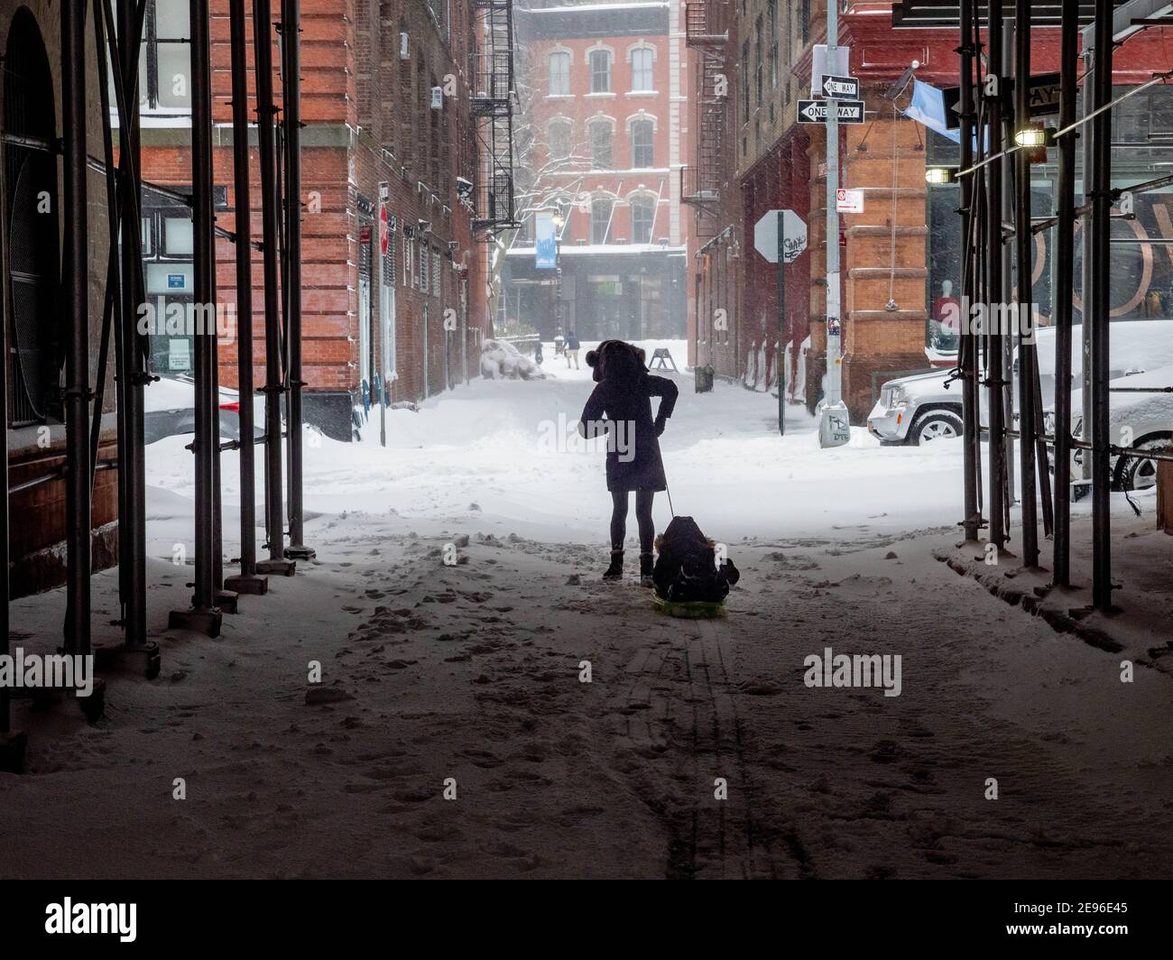 New York, New York, USA. Februar 2021. Eine Mutter zieht ihren Sohn auf einem Schlitten durch die Staple Street in Tribeca. Diese ikonische NYC Straße mit ihrer Skybridge, die jetzt von Gerüsten umschlossen ist, wurde in unzähligen Filmen und tv-Shows gezeigt. Quelle: Milo Hess/ZUMA Wire/Alamy Live News Stockfoto