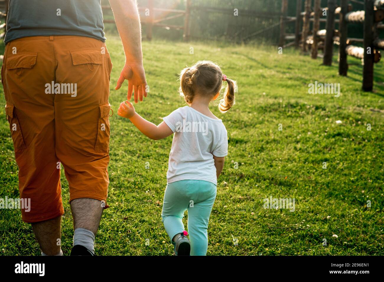 Ein Elternteil hält die Hand eines kleinen Kindes.Baby Mädchen geht Hand in Hand mit Papa im Dorf bei Sonnenuntergang. Sonniger Abend Stockfoto