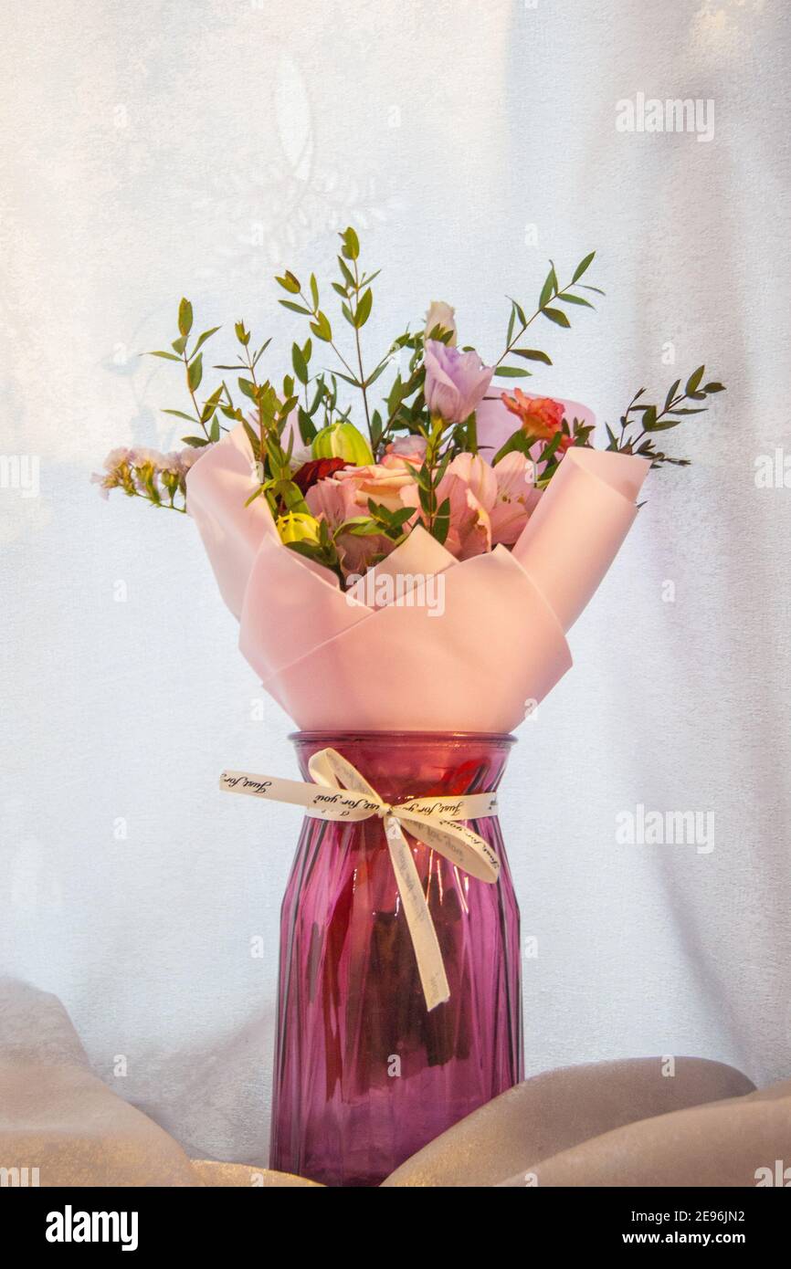 Ein zartes Bouquet aus verschiedenen Blumen in einer hellrosa Umhüllung steht auf einem hellen Hintergrund in einer häuslichen Umgebung. Vertikales Foto. Stockfoto