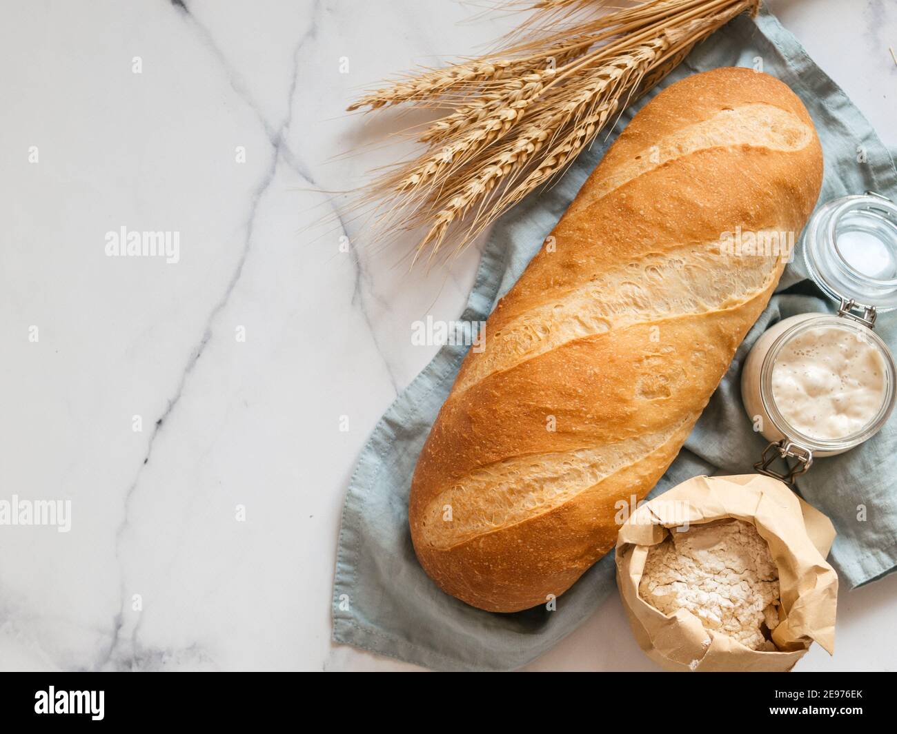 Britisches Weißblüher oder europäischer Sauerteig Baton-Brot auf weißem Marmorgrund. Frisches Brot und Glas mit Sauerteig Starter, Mehl in Papiertüte und Ohren. Draufsicht. Kopierbereich Stockfoto