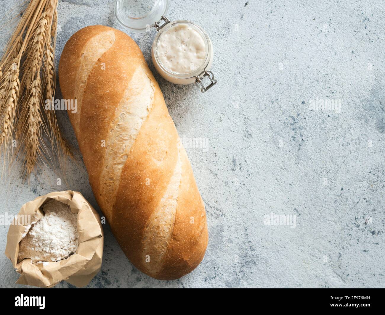 Britisches Weißblüher oder europäischer Sauerteig Baton-Brot auf grauem Zementgrund. Frisches Brot und Glas mit Sauerteig Starter, Mehl in Papiertüte und Ohren. Draufsicht. Kopierbereich Stockfoto