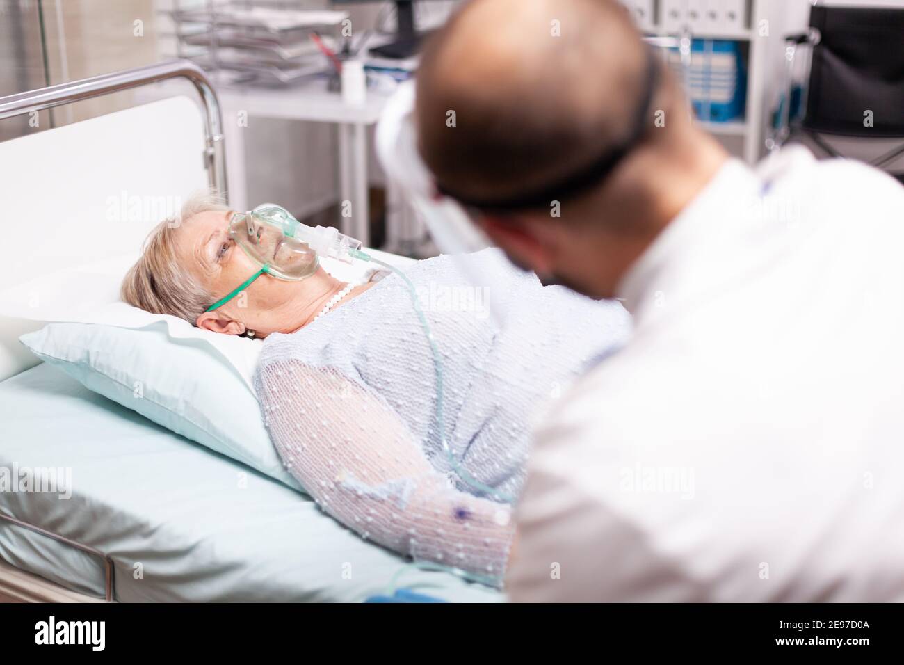 Ältere Frau sitzt auf Klinikbett mit Sauerstoffmaske in der Zeit von covid19. Patient im Krankenhauszimmer, der während einer Coronavirus-Pandemie auf einem medizinischen Bett mit infizierter Lunge liegt und ein Atemproblem hat. Stockfoto