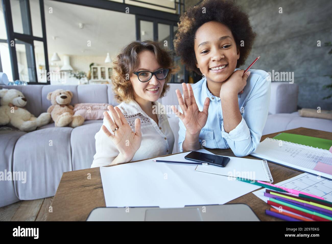 Freudige gemischte Rasse teen Mädchen und weibliche Lehrer winken an Kamera während Hausaufgaben zusammen im Wohnzimmer zu tun Zu Hause Stockfoto