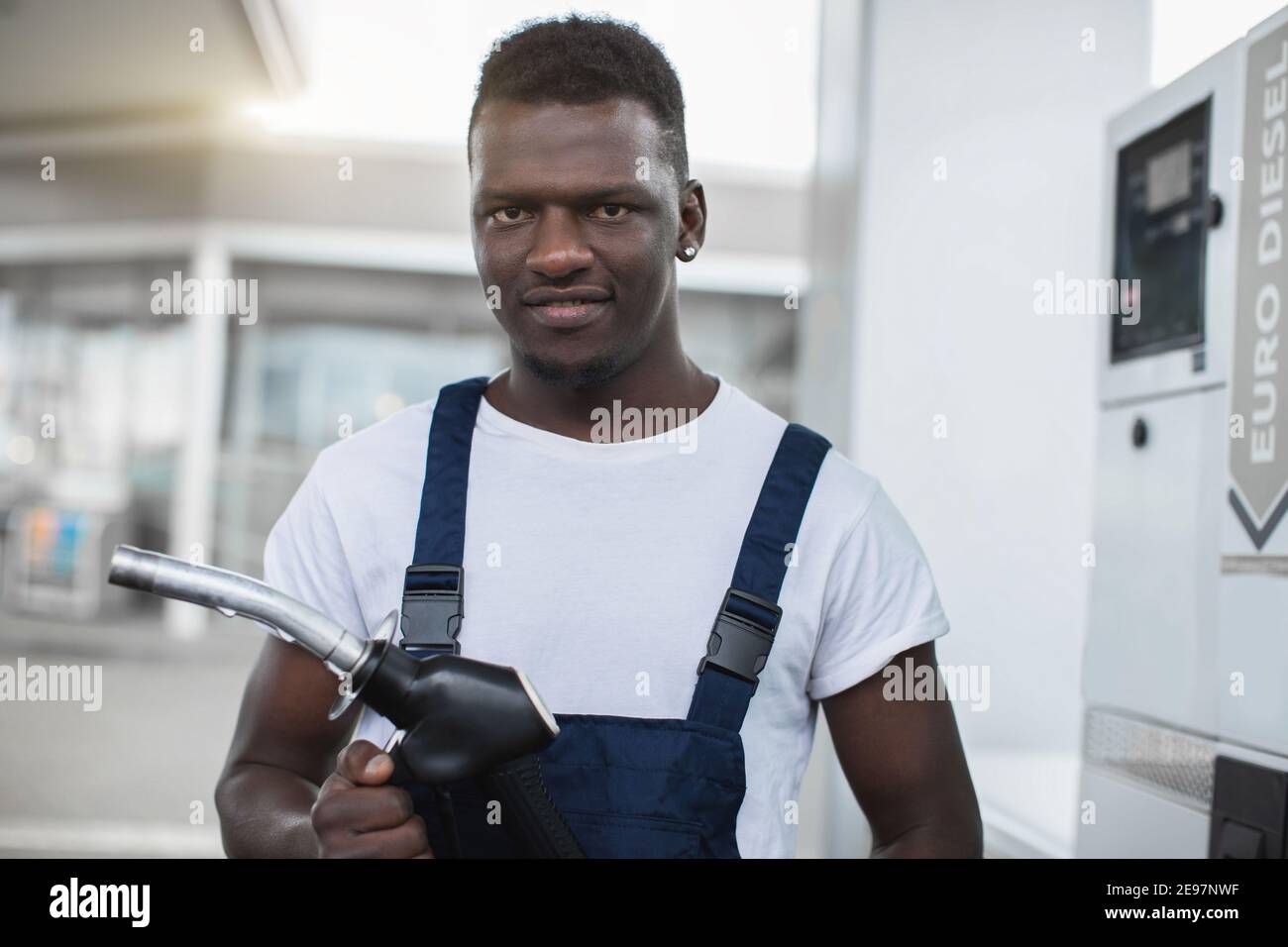 Nahaufnahme eines Tankstellenarbeiters, eines hübschen jungen afroamerikanischen Mannes, der weißes T-Shirt und blaue Overalls trägt und mit einem Lächeln vor der Kamera posiert Stockfoto