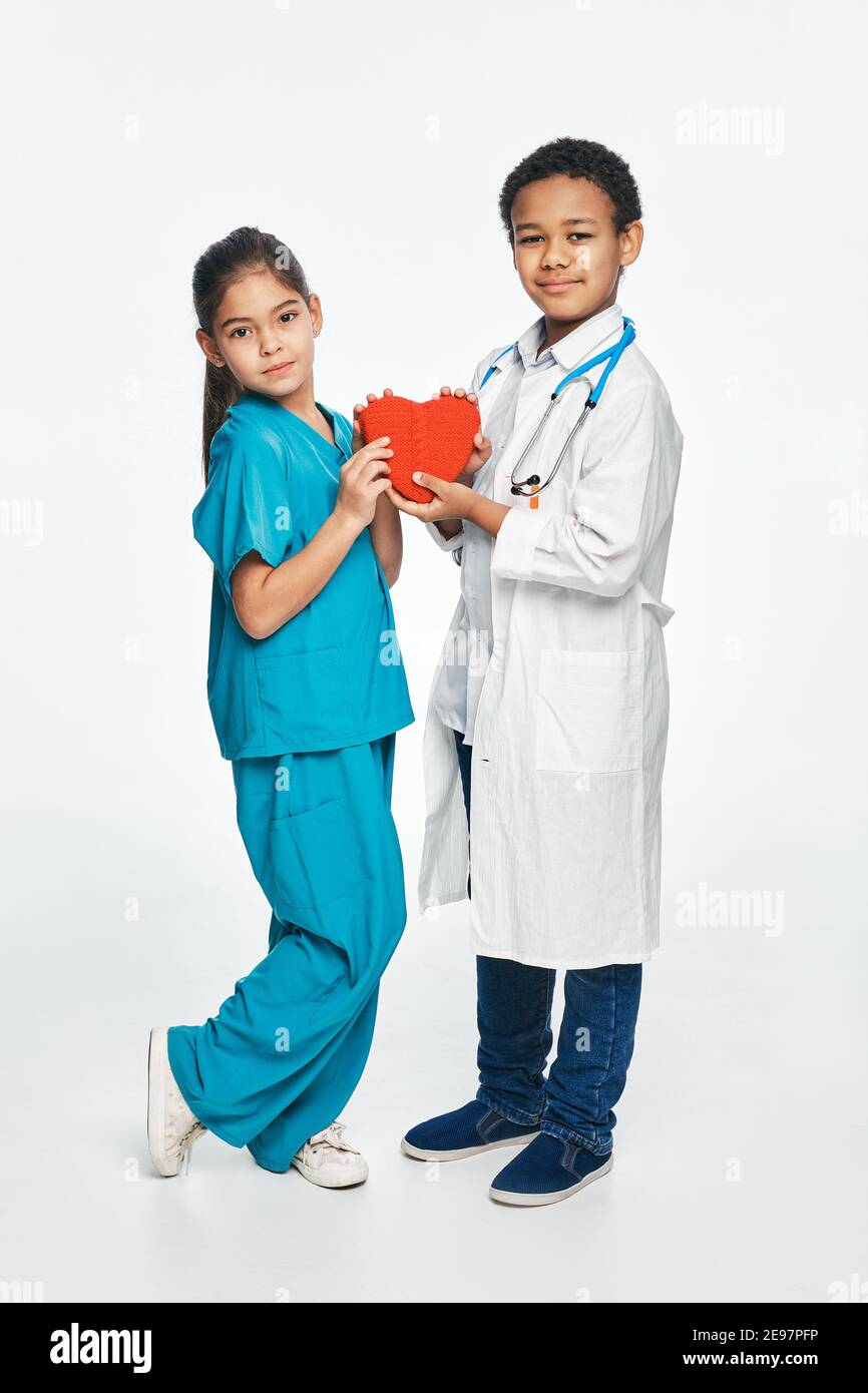 Gruppe von multiethnischen Kindern tragen medizinische Uniformen, halten Kinder Form des Herzens in den Händen. Zukünftige Herzärzte Stockfoto