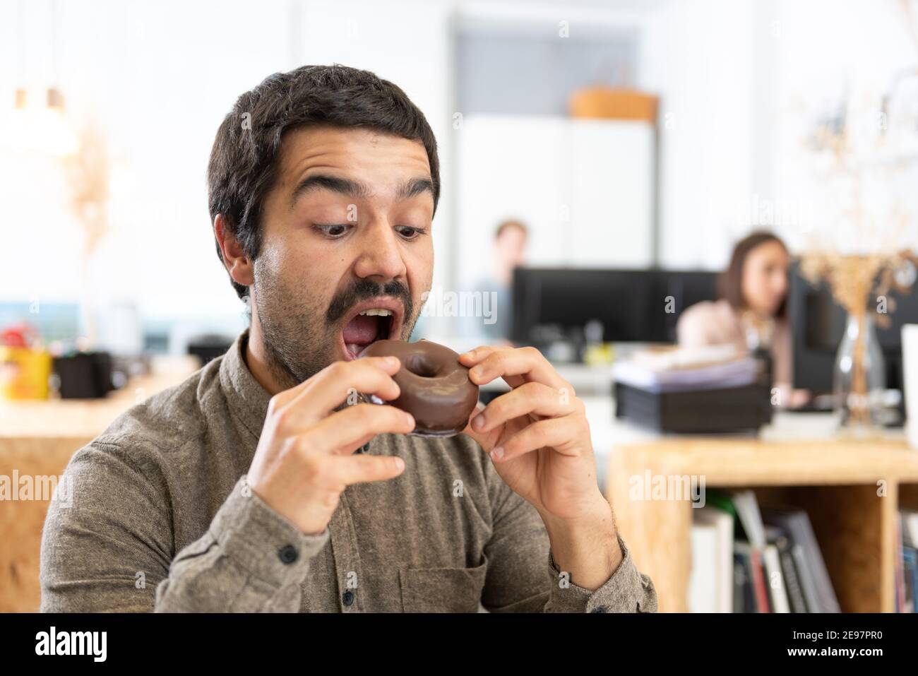 Hispanic Mann mit Schnurrbart essen ein Schokolade süßen Brötchen. Ungesunde Gewohnheiten im Bürokonzept. Stockfoto
