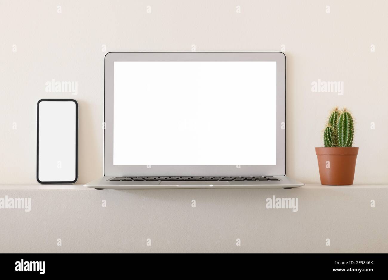 Smartphone und Laptop mit weißen leeren Bildschirmen mit Topflastern platziert Kaktus auf Regal gegen helle Wand Stockfoto