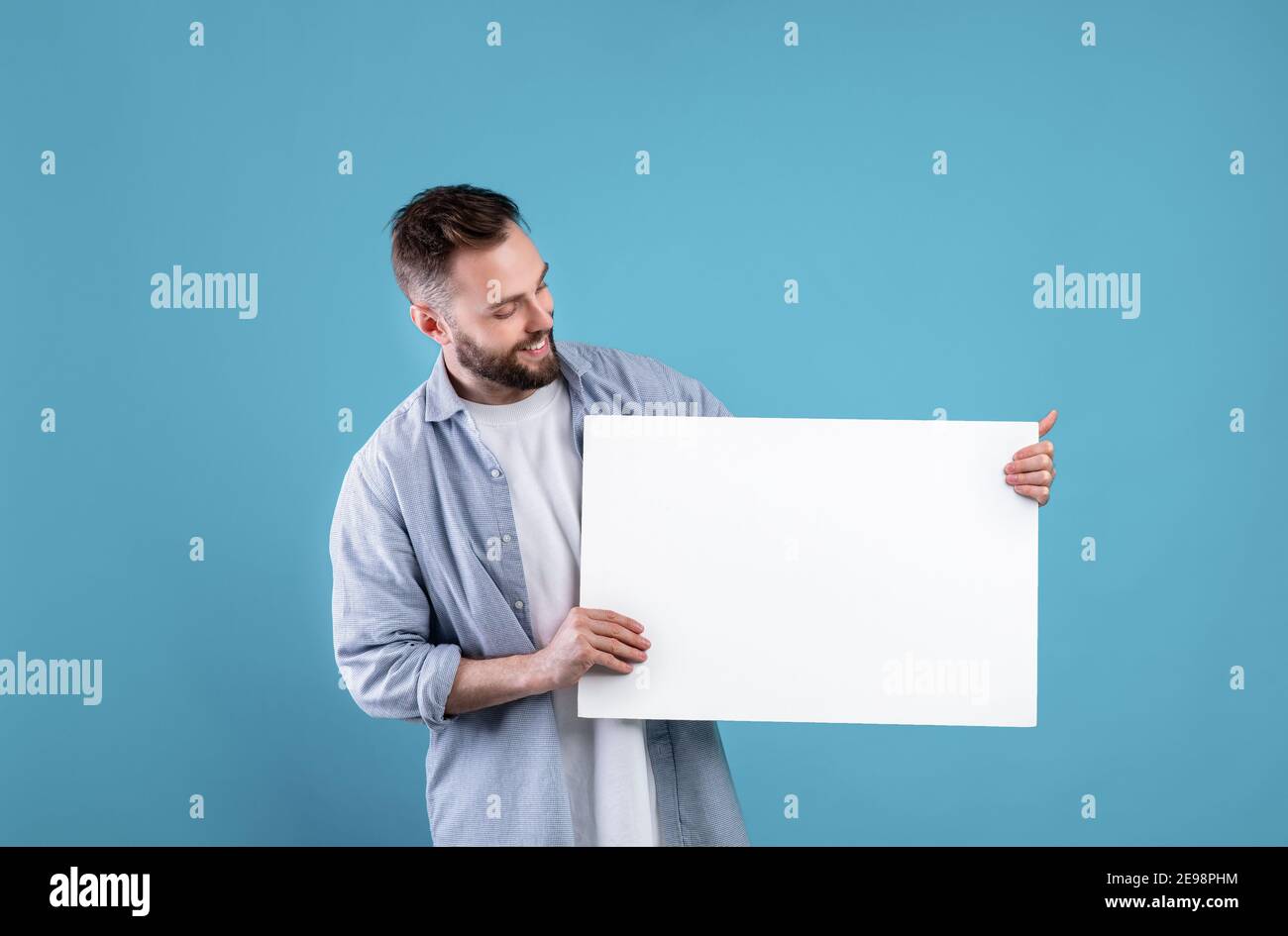 Glücklich lächelnder Mann, der weiße leere Plakette auf blauem Studiohintergrund anzeigt, mockup für Logo oder Design Stockfoto
