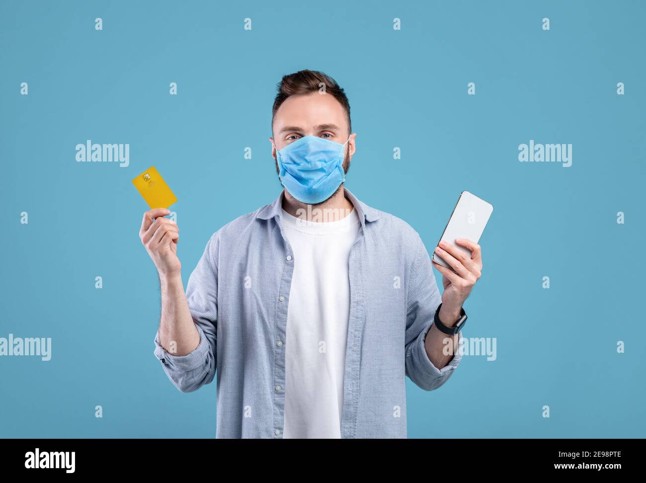 Kontaktloses Einkaufen während der covid Quarantäne. Junger Mann in Maske hält Handy und Kreditkarte, blauer Hintergrund Stockfoto