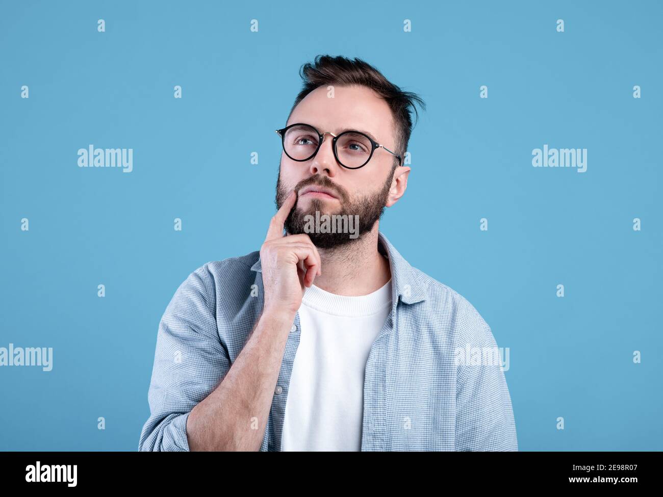 Porträt eines nachdenklichen bärtigen Mannes in einer Brille, der sein Kinn berührt, tief im Gedanken über blauen Studiohintergrund Stockfoto