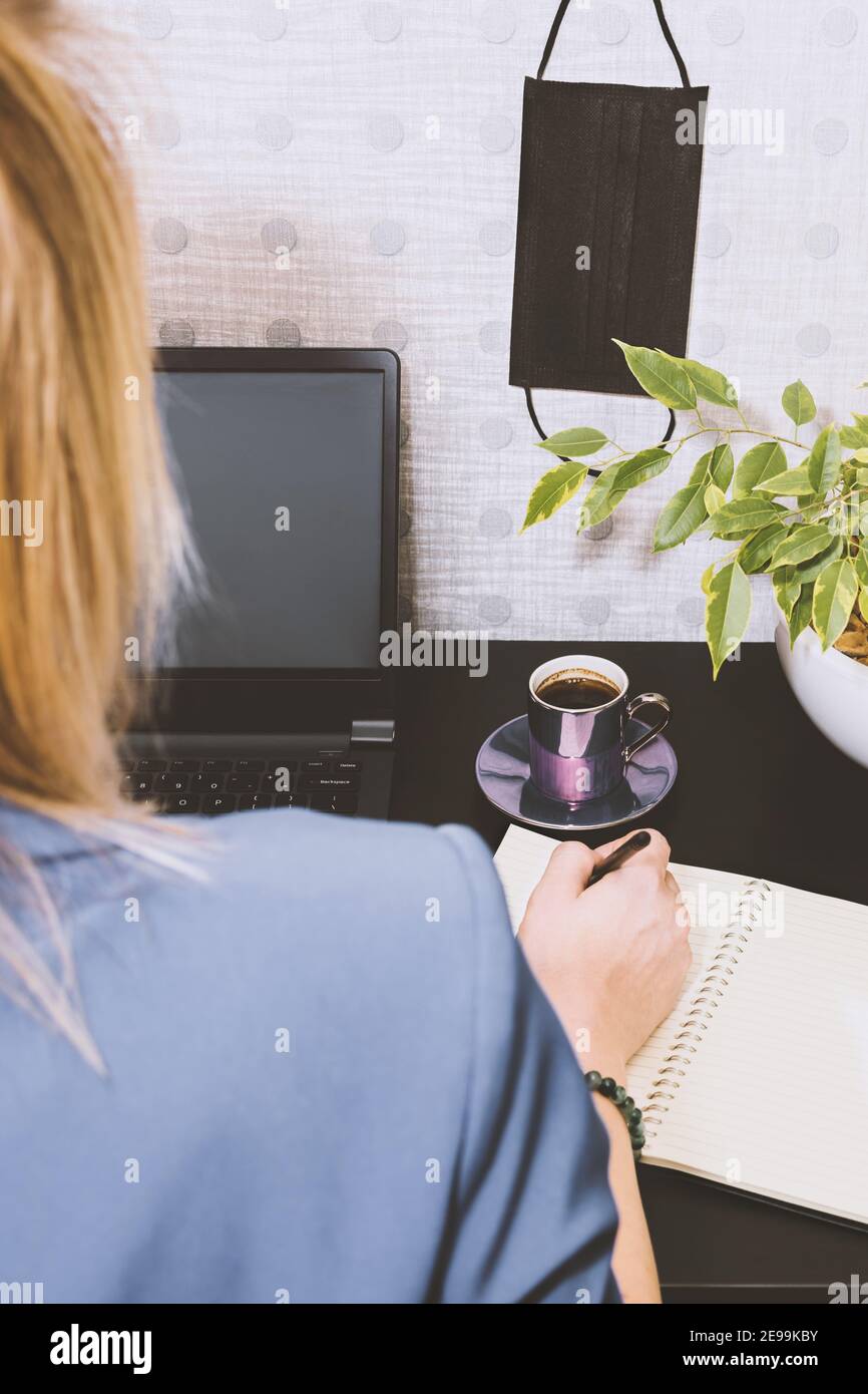 Gesundheitskonzept. Blonde Frau arbeitet mit Computer von zu Hause aus während Quarantäne und sitzt am Schreibtisch mit Notizen, Kaffeetasse, grüne Blume und ha Stockfoto