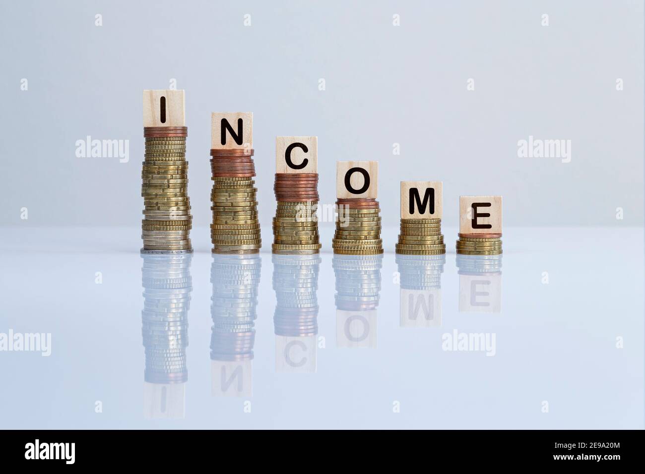 Wort "EINKOMMEN" auf Holzblöcken auf absteigenden Münzstapeln auf grauem Hintergrund. Konzeptfoto von Geldverlust, Wirtschaftskrise und Rezession. Stockfoto