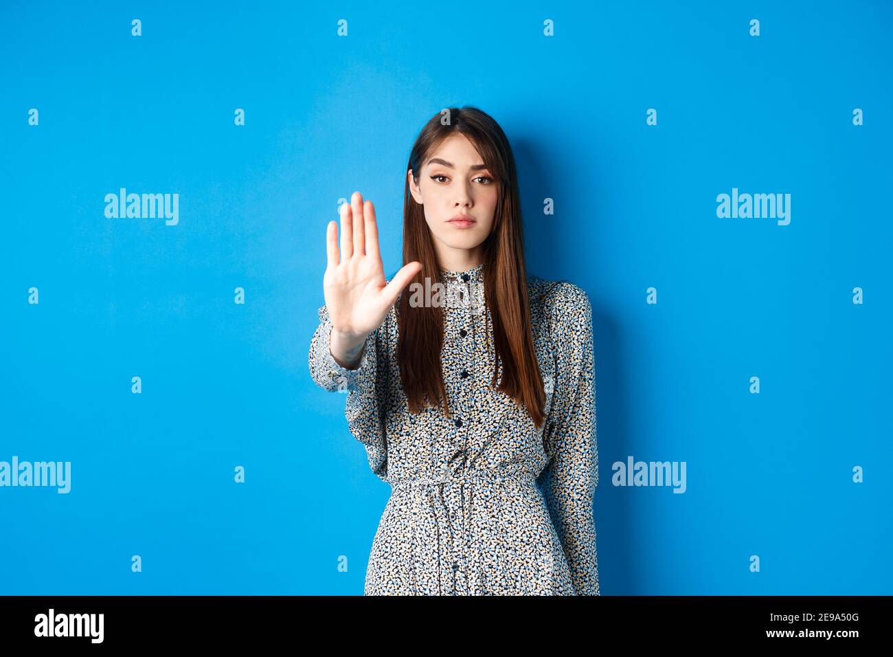 Seriöse und selbstbewusste Frau mit langen Haaren und Kleid, Strecke die Hand aus und sage nein, machen Stop-Geste, verbieten schlechte Aktion, stehen auf blauem Hintergrund Stockfoto