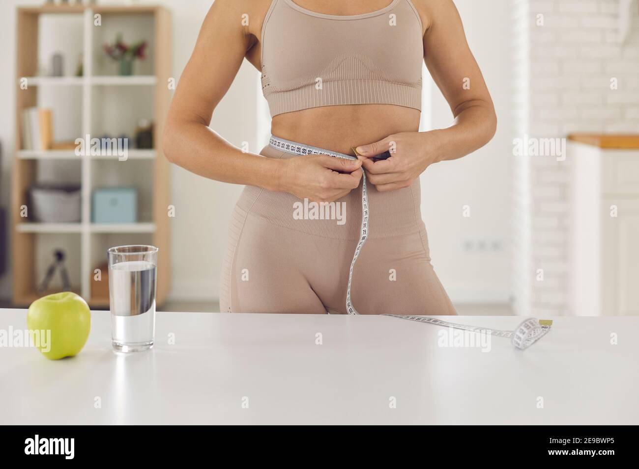 Abgeschnittene Aufnahme von fit Frau Messung Taille stehen am Tisch Mit Apfel und Glas Wasser Stockfoto