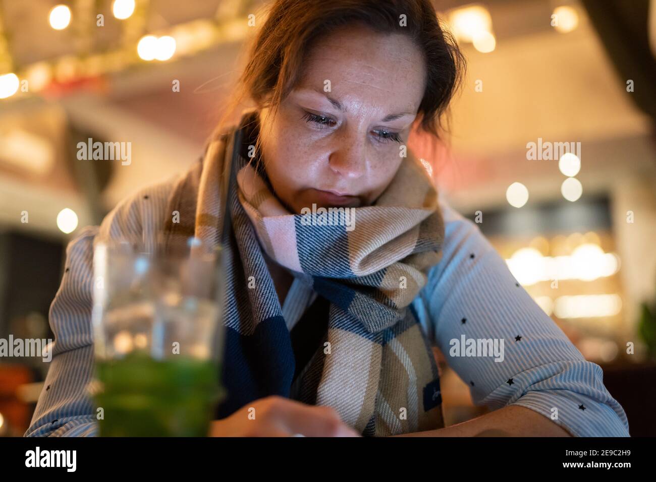 Junge Frau trägt Schal mit Getränk in hellen bunten Cocktail Bar mit Handy Blick auf den Bildschirm Beleuchtung ihr Gesicht vom Telefon Stockfoto