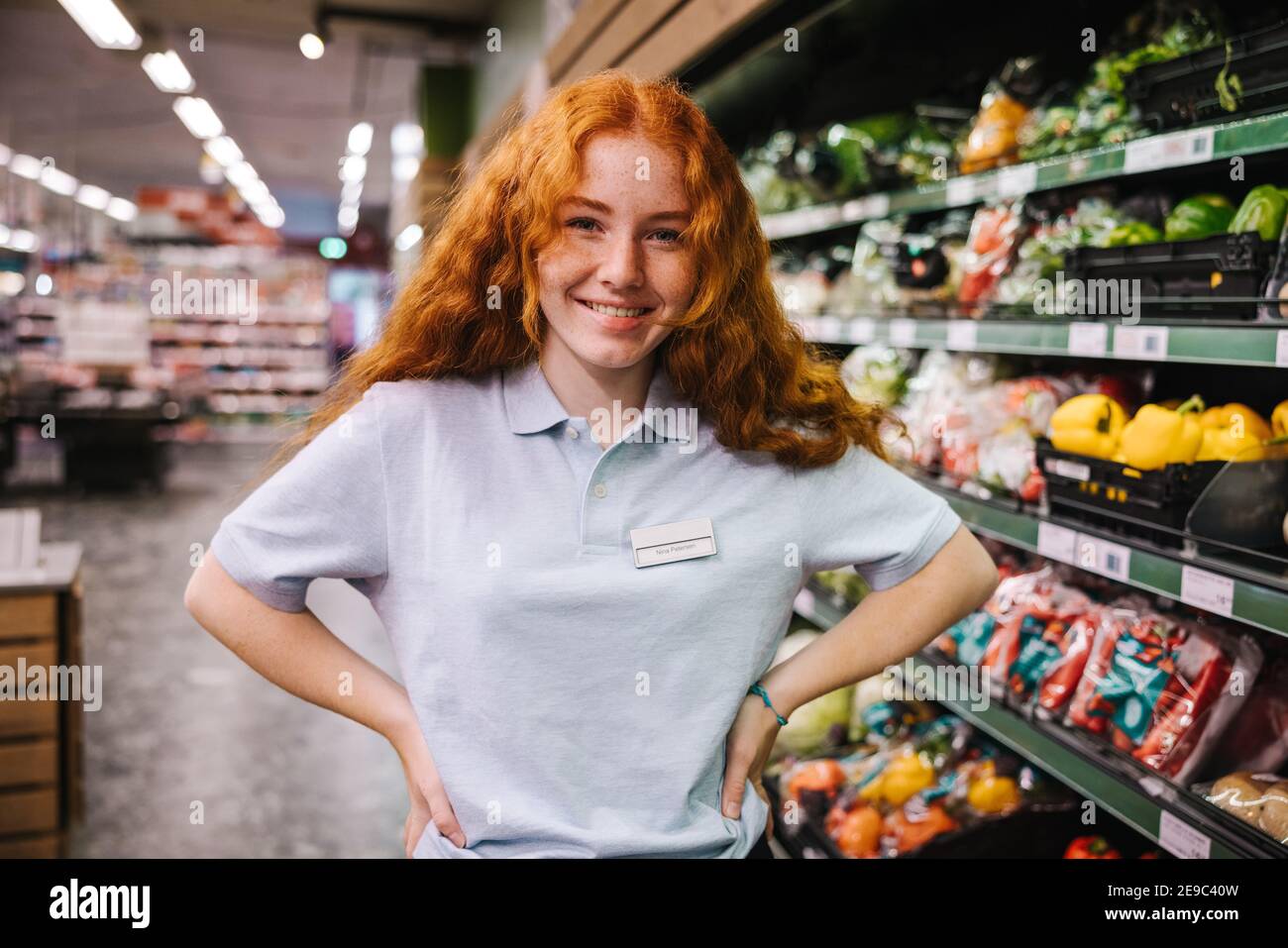 Porträt einer Arbeiterin in einem Lebensmittelgeschäft und lächelnd auf die Kamera schauend. Frau in Uniform stehend mit Händen auf Hüften im Supermarkt. Stockfoto