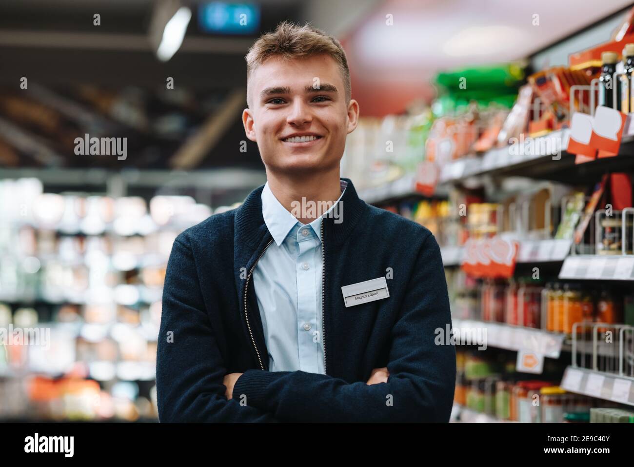 Porträt eines jungen Arbeiters in einem Lebensmittelgeschäft und Blick auf die Kamera lächelnd. Mann in Uniform stehend mit gekreuzten Armen im Supermarkt. Stockfoto