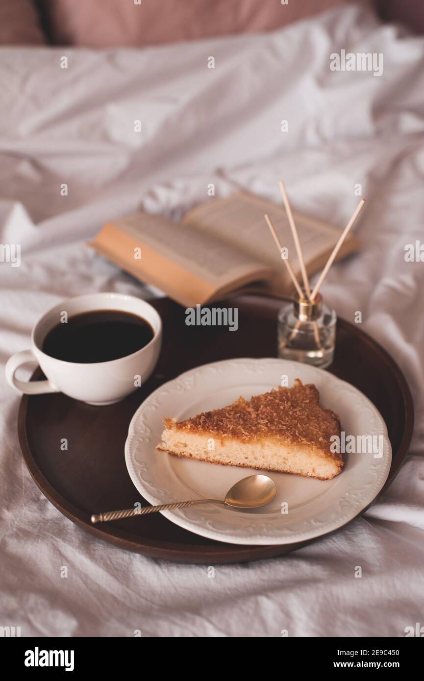 Tasse frischen Kaffee mit leckeren Kuchen auf weißem Teller mit offenem Buch im Bett über strahlende Lichter aus nächster Nähe. Guten Morgen. Frühstückszeit. Stockfoto