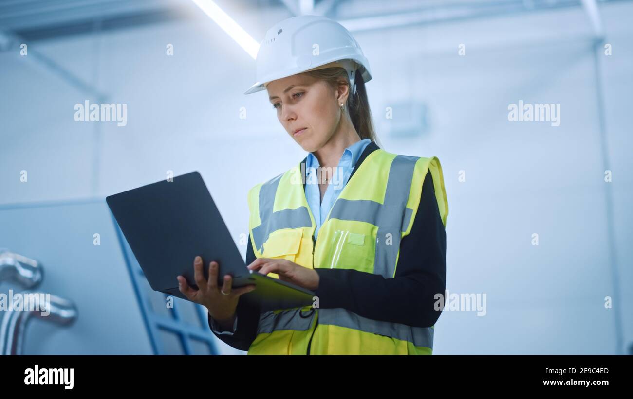 High-Tech-Fabrik: Selbstbewusste und professionelle weibliche Ingenieurin trägt Sicherheitsjacke und Harthut Halten und Arbeiten auf Laptop-Computer. Modern Hell Stockfoto