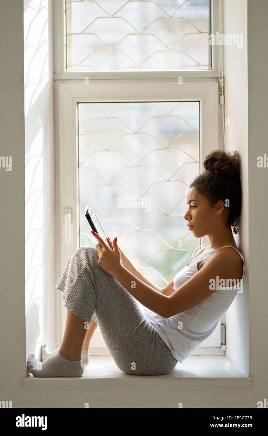 Seitenansicht einer entspannten jungen Frau, die mit einem Tablet-pc ein Buch liest, während sie zu Hause am Fenster sitzt. Lifestyle, Freizeitkonzept. Seitenansicht Stockfoto Seitenansicht einer entspannten jungen Frau, die mit einem Tablet-pc ein Buch liest, während sie zu Hause am Fenster sitzt. Lifestyle, Freizeitkonzept. Seitenansicht Stockfoto