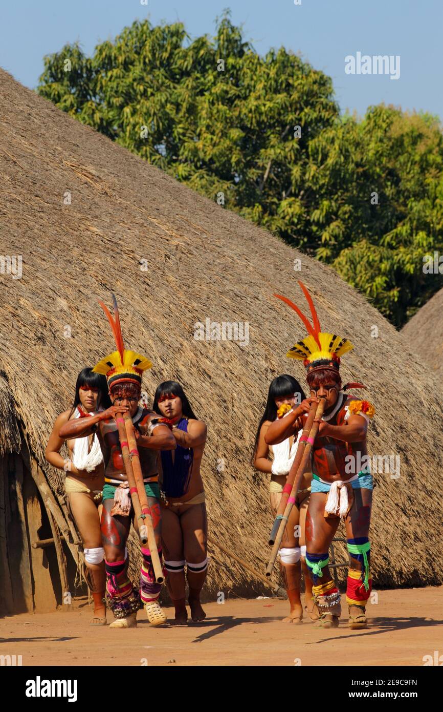 Zwei einheimische Indios mit zwei jungen Mädchen in festlicher Kleidung und Kunststücken im Haar, die ihre lange Pfeife spielen, Mato Grosso, Brasilien, Südamerika. Stockfoto