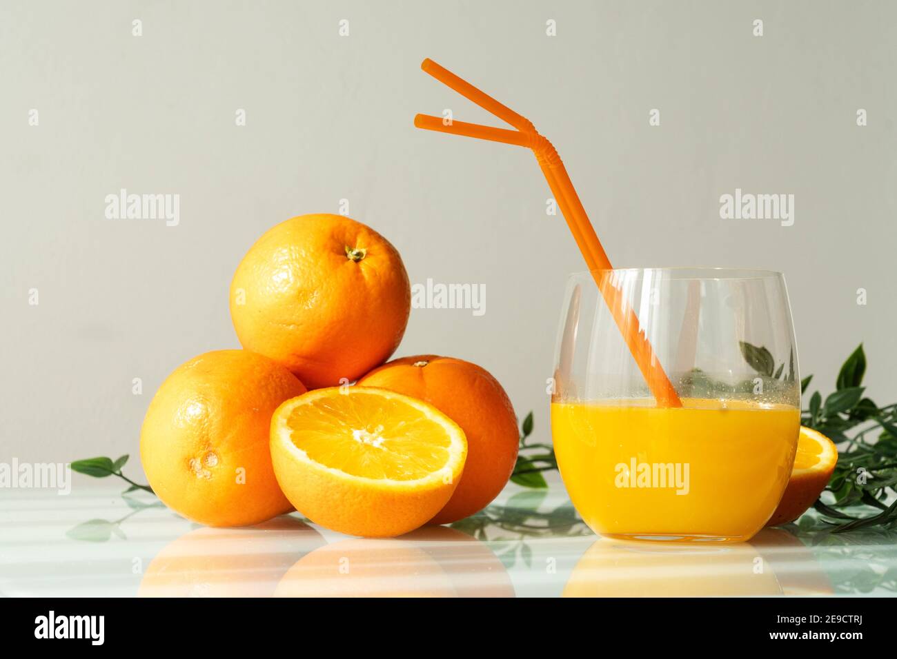 Orangensaft in einem Glas, umgeben von natürlichen Orangen und einigen grünen Blättern, mit einem natürlichen Sonnenlicht hellweißen Hintergrund Stockfoto