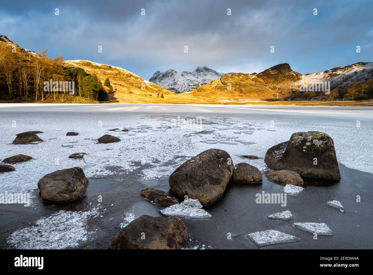 Gefrorener See mit Felsen und Eis an der Küste. Blick auf schneebedeckte Berge mit goldenem Morgenlicht. Blea Tarn, Lake District, Großbritannien. Stockfoto