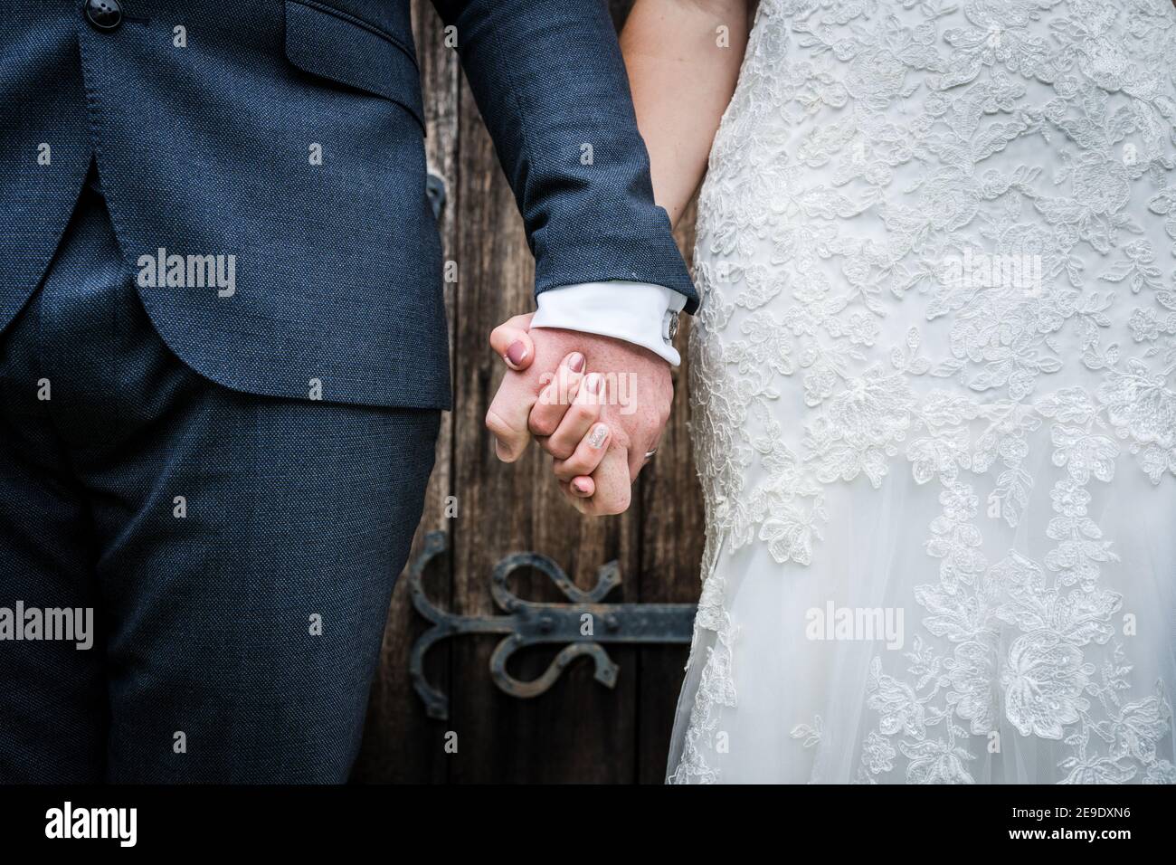 Nahaufnahme der Braut in weißem Kleid und Bräutigam in Anzug gerade verheiratet hält Hände zusammen nach der Hochzeitszeremonie stand Vor der hölzernen Kirchentür Stockfoto