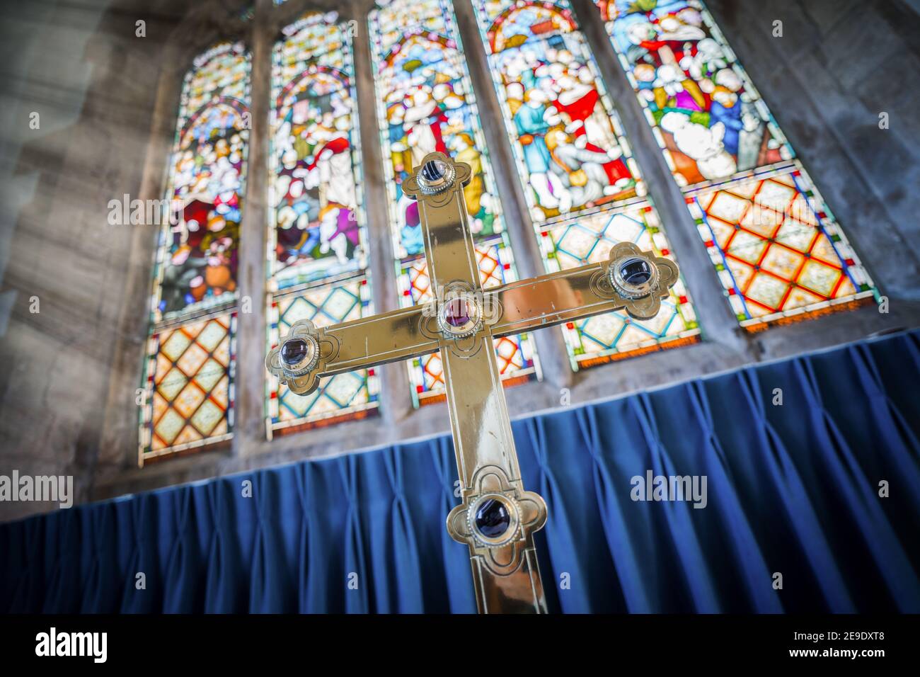 Großes Messinggoldenkreuz mit funkelnden Juwelen stand im Sonne vor der Kirche von England Kapelle mit großen Hohe bunte Buntglasfenster Stockfoto