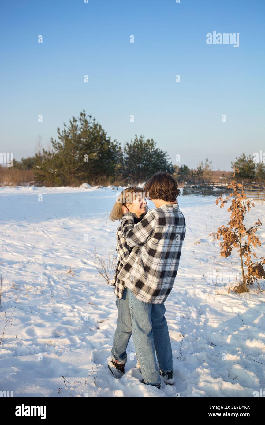 Fröhliches Teenager-Mädchen und Junge, etwa 16-17 Jahre alt, in karierten Hemden und Jeans, stehen im Schnee umarmt und lachen fröhlich und schauen einander an. Stockfoto