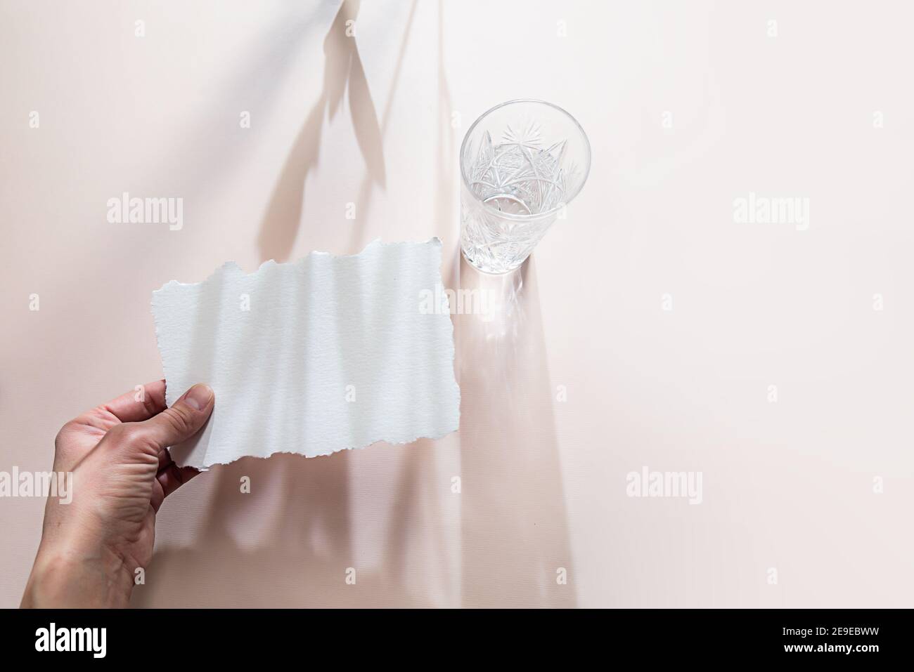 Minimalistisches Stillleben im Sommer. Eine weibliche Hand hält ein weißes leeres Blatt Papier, ein Glas Wasser steht auf einem hellen Tisch, lange Schatten, Strahlen der Stockfoto