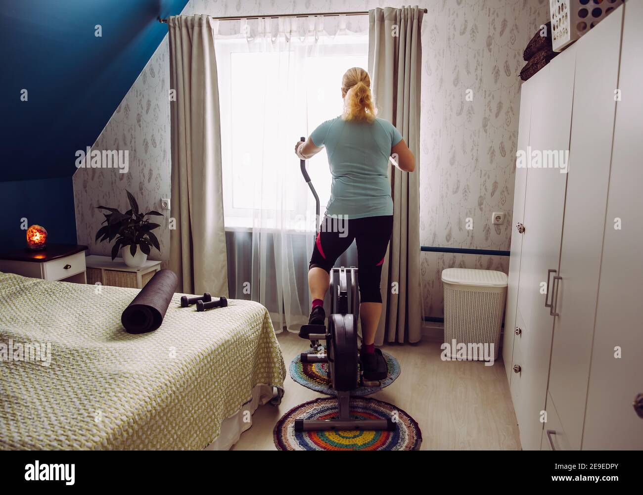 Übergewichtige Frau tun Training mit Crosstrainer zu Hause während der Quarantäne und schauen aus dem Fenster. Zeit für Selbstverbesserungskonzept. Stockfoto