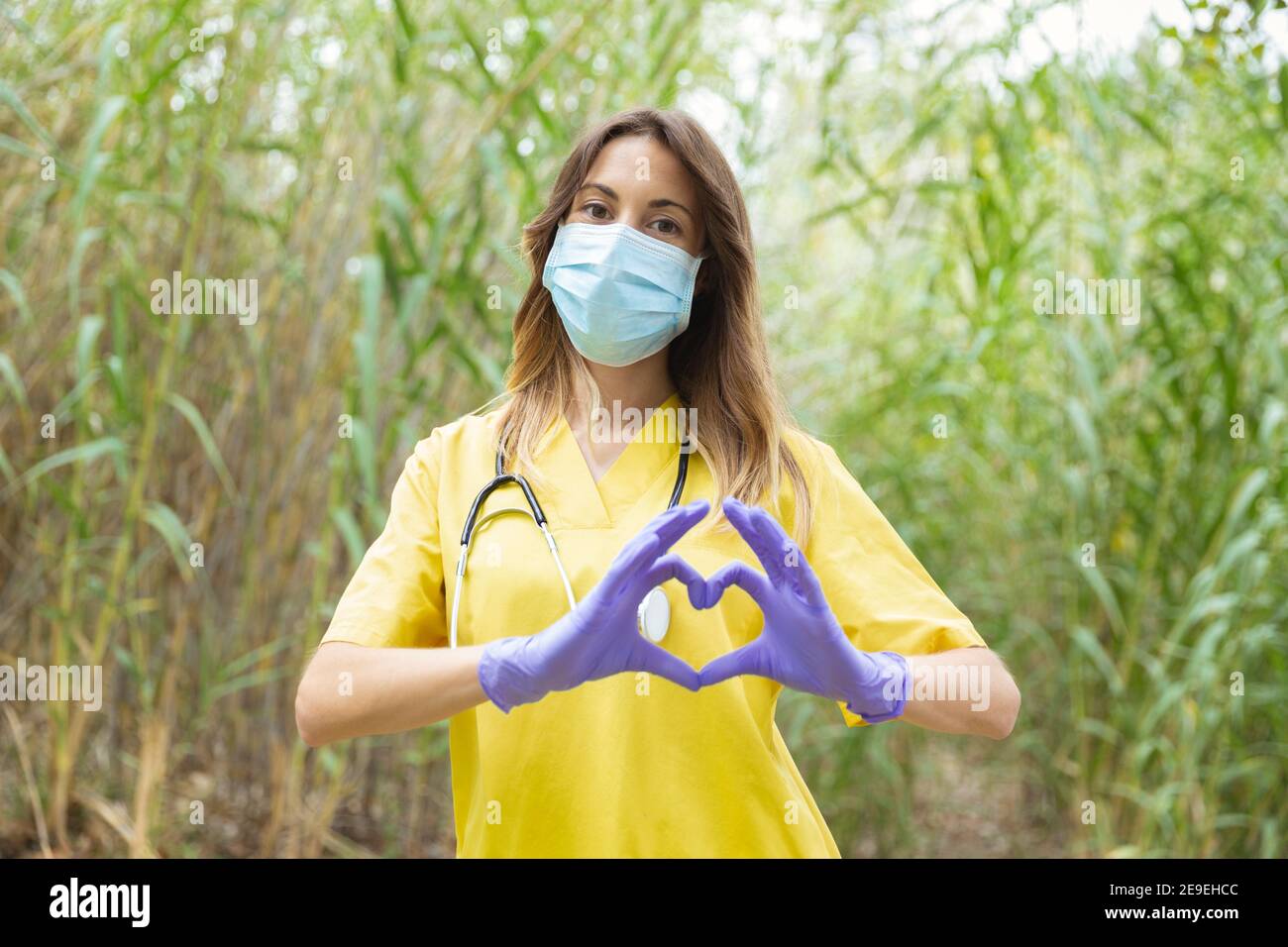 Kaukasische Krankenschwester in Uniform, Handschuhe und OP-Maske, bilden ein Herz mit ihren Händen. Sie ist im Freien in der Natur. Leerzeichen für Text. Stockfoto