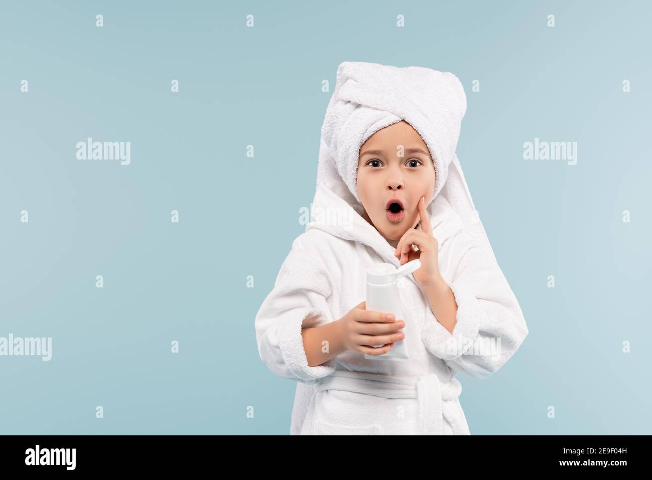 Schockiert Kind in Bademantel und Handtuch auf Kopf halten Rohr Mit kosmetischem Produkt isoliert auf blau Stockfoto