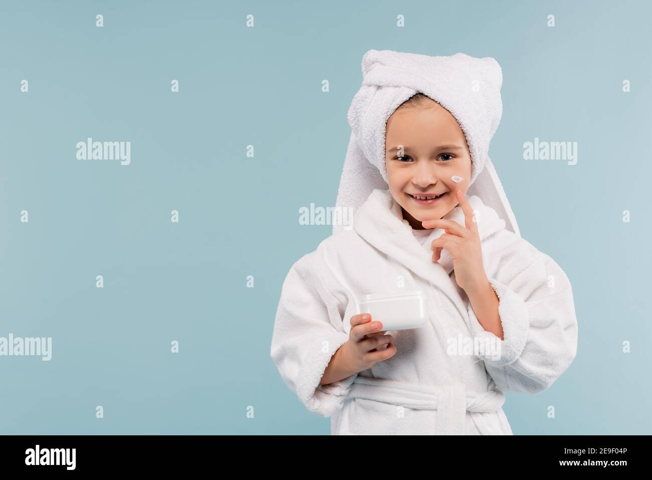 Fröhliches Kind im Bademantel und Handtuch auf dem Kopf halten Behälter Und Anwendung kosmetische Produkt isoliert auf blau Stockfoto